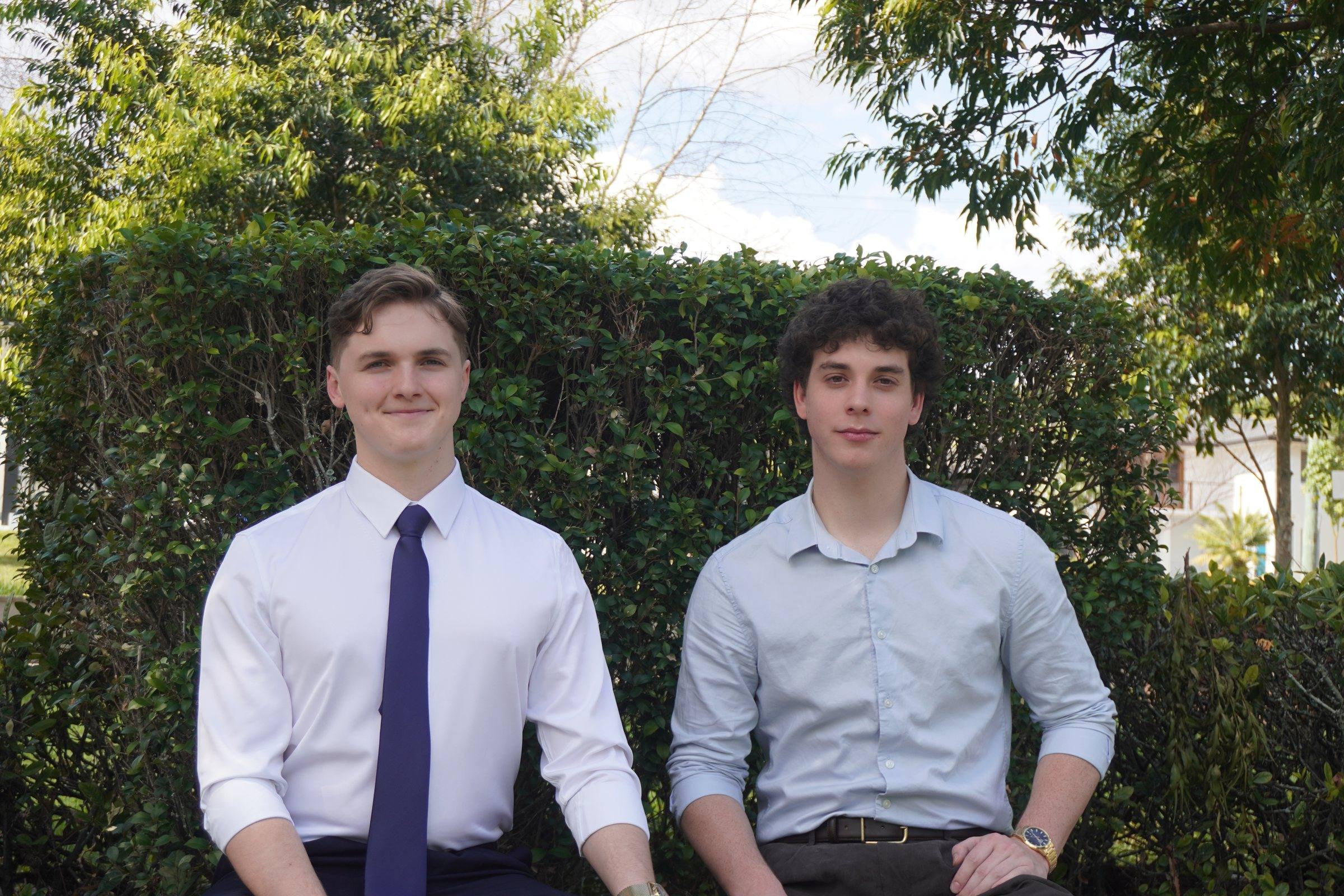 Zeek and Levi sitting side by side in front of a tall green hedge