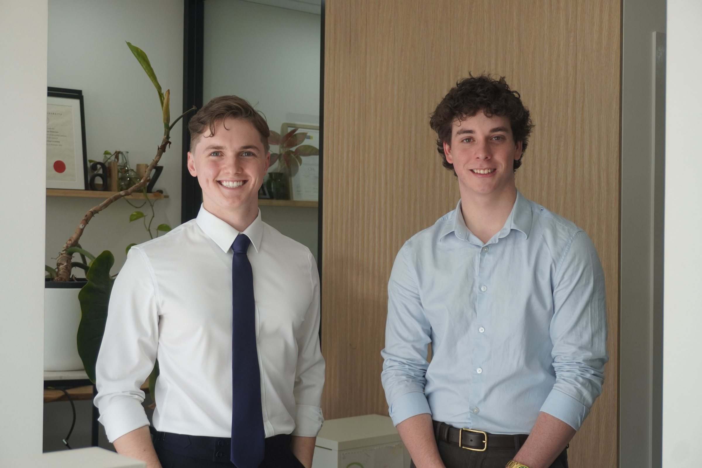 Zeek in a white shirt and tie and Levi in a light-blue shirt, standing together in an office, smiling